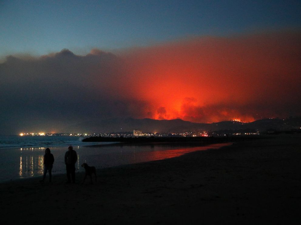 Two people walk along the beach with a dog as a wildfire burns in distance Tuesday, Dec. 5, 2017, in Ventura, Calif. Raked by ferocious Santa Ana winds, explosive wildfires northwest of Los Angeles and in the citys foothills burned a psychiatric hos